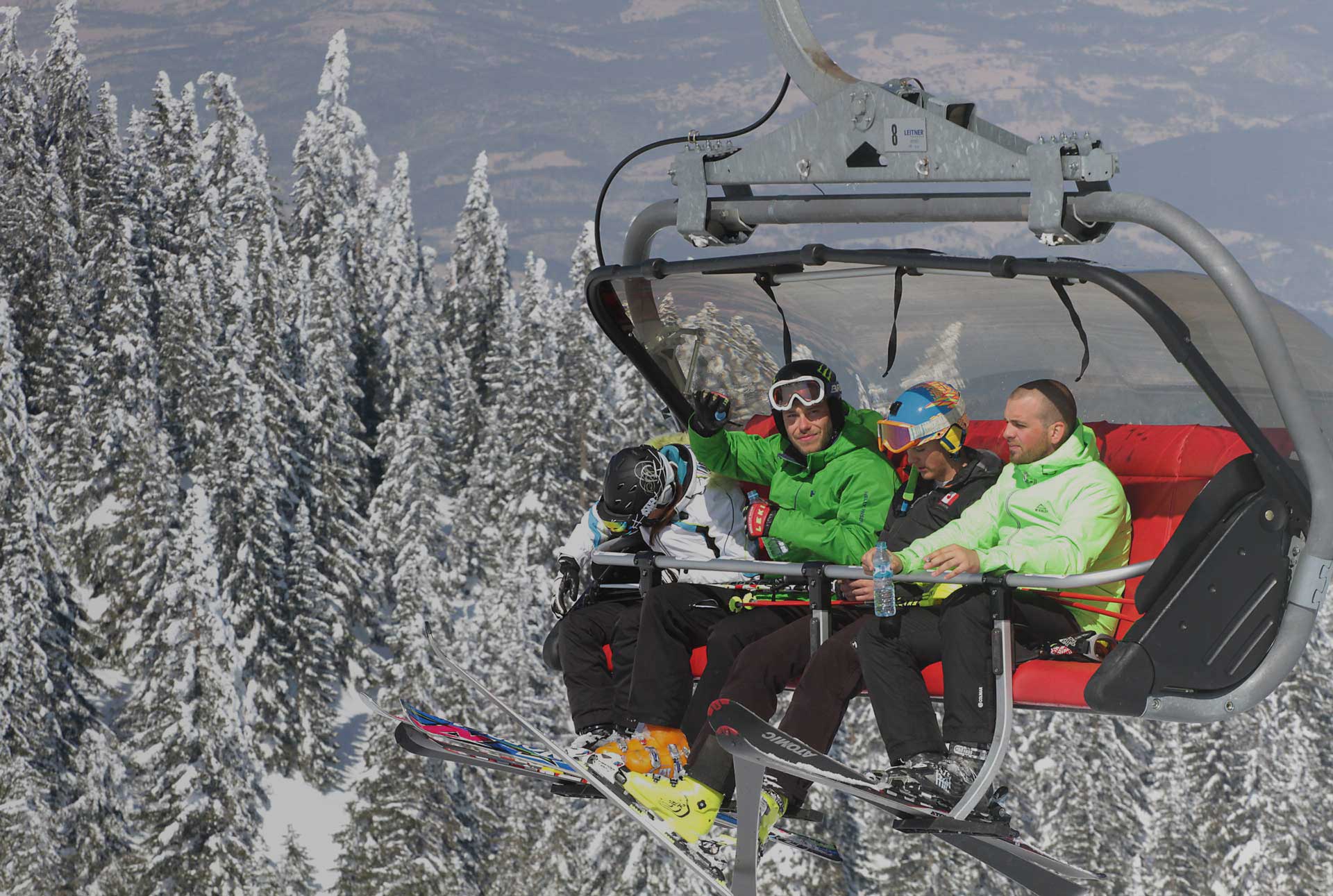 The image shows four skiers riding a chairlift up a snowy mountain. They are comfortably seated, their skis hanging below, and are surrounded by a picturesque view of snow-covered evergreen trees. One skier is waving, suggesting a feeling of joy and exhilaration associated with the winter sport. The overall mood is one of fun, adventure, and the beauty of a winter landscape.