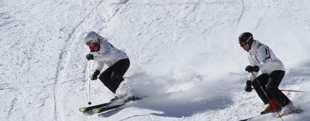 The image shows two skiers, a man and a woman, in white ski suits, skiing down a snowy slope.